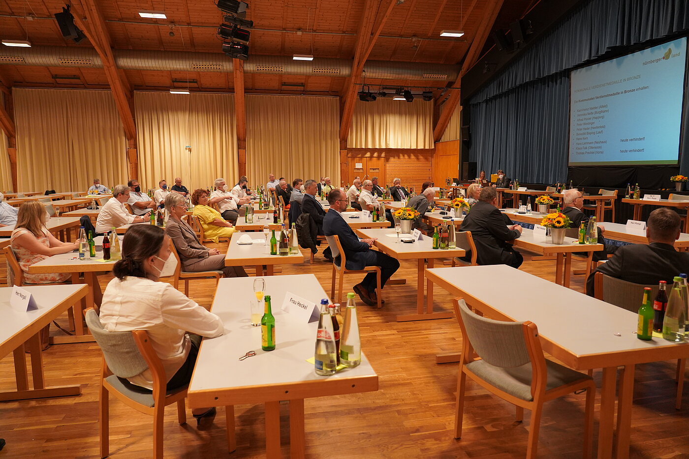 In etwa 30 Menschen unterschiedlichen Alters und Geschlechts sitzen an Tischen in der Karl-Diehl-Halle in Röthenbach und schauen zur Bühne. Dort gibt es auf einer großen Leinwand eine Präsentation zu sehen.